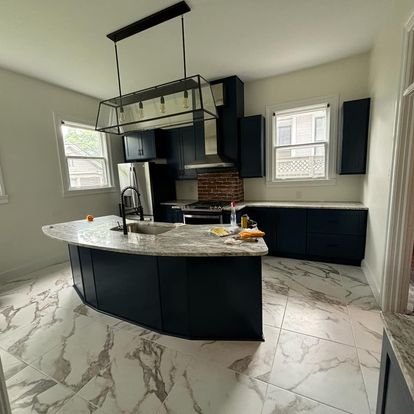 A modern kitchen featuring dark blue cabinets, a large island with a marble countertop, and large windows allowing natural light. The kitchen has stainless steel appliances and a brick accent wall.