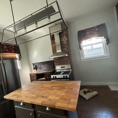 A cozy kitchen with a wooden countertop island, brick wall, and stainless steel appliances. The kitchen has soft lighting and window treatments that add charm.