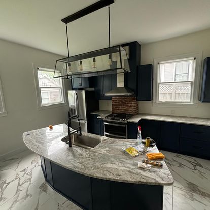 A stylish kitchen with dark blue cabinetry, marble countertops, and ample natural light from nearby windows. The space includes stainless steel appliances and a light fixture above the island.