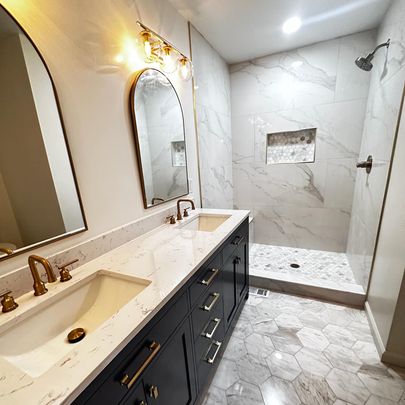 An elegant bathroom with dual sinks, gold fixtures, and a tiled shower area with two mirrors reflecting the stylish decor.