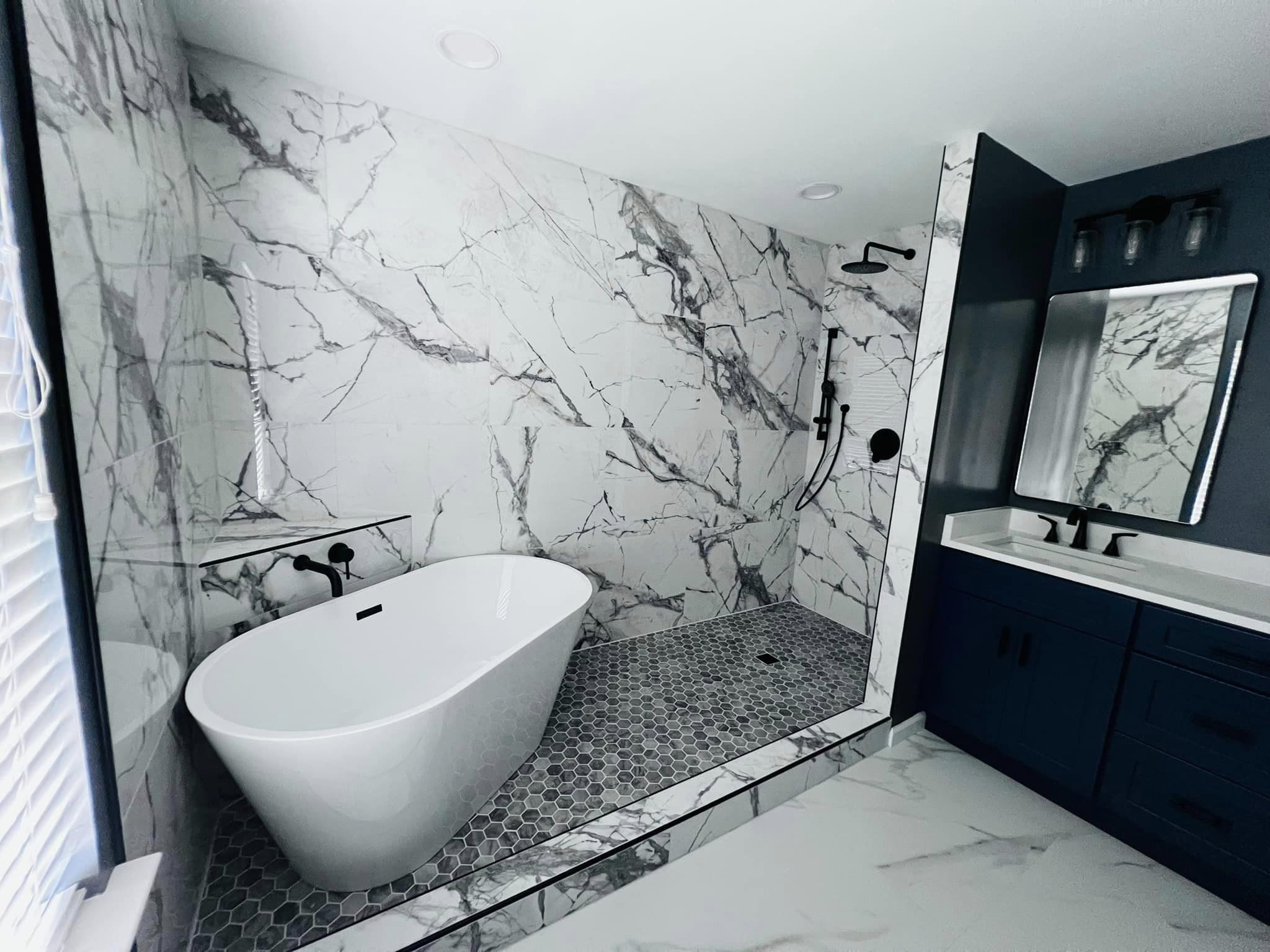 A modern bathroom remodel with a white bathtub, a walk-in shower, and a blue vanity. The walls are tiled with white marble, and the floor is tiled with grey hexagonal tiles.