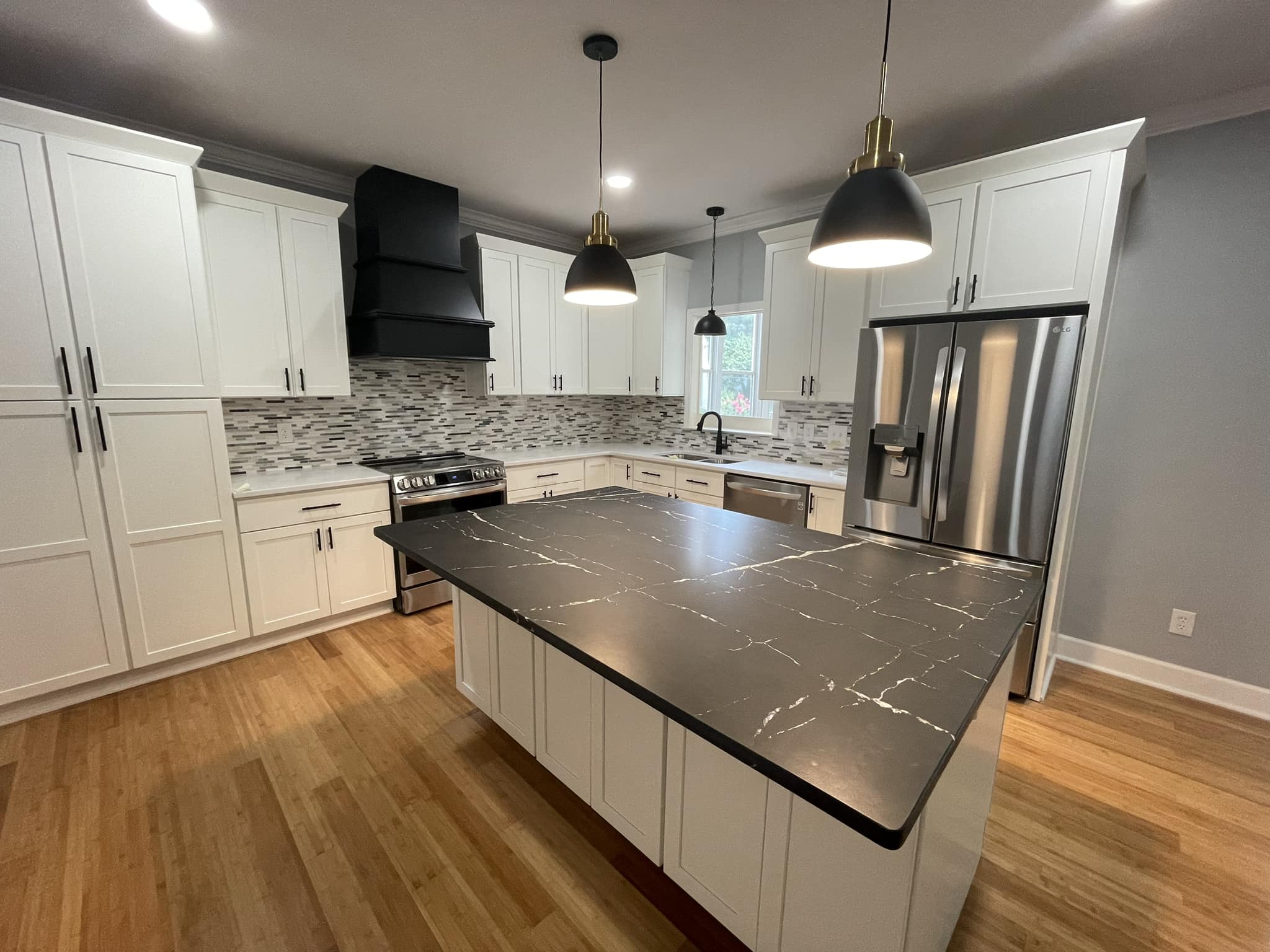 A contemporary kitchen with white cabinets and a striking black countertop island. It features modern lighting, glass tile backsplash, and stainless steel appliances.