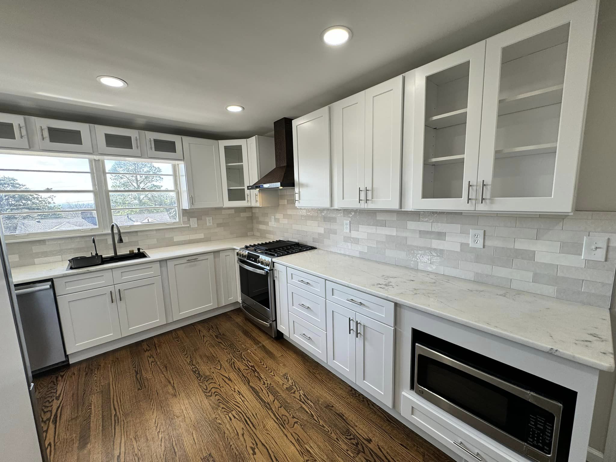 A bright kitchen with white cabinetry, stainless steel appliances, and a marble countertop. The setup includes a large window and a mixture of drawer and cupboard storage.